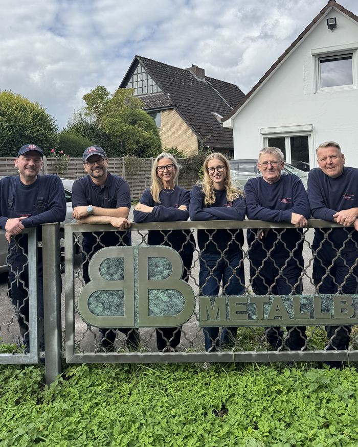 Das Team der Bruhn & Beilfuss GmbH lehnt lachend an einem Metallzaun mit BB-Emblem