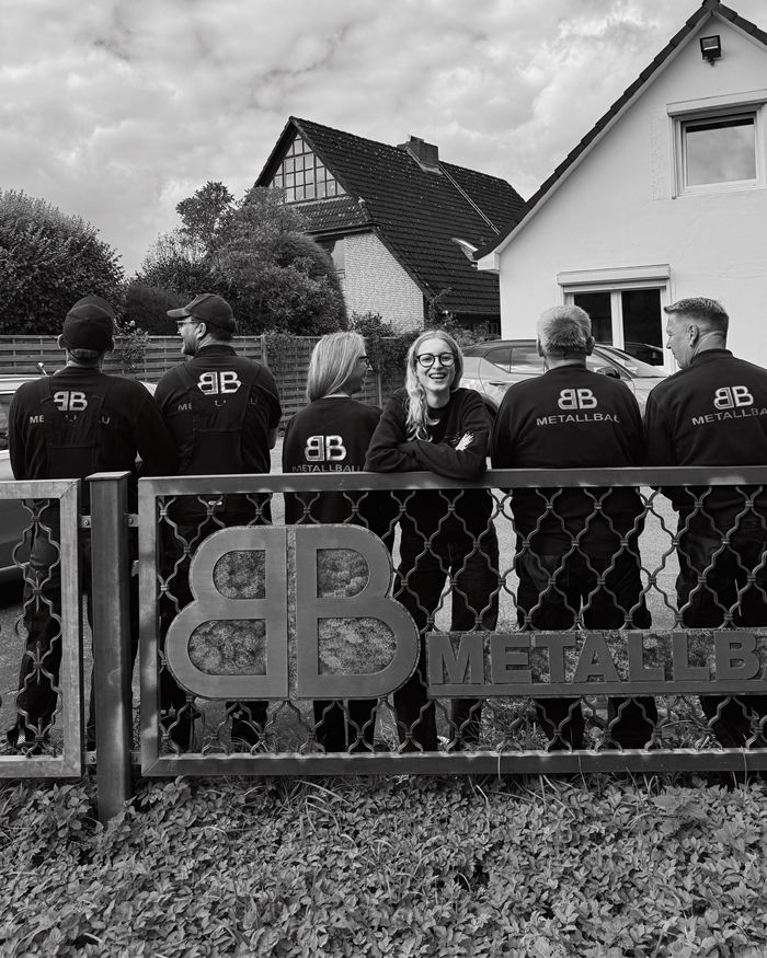 Schwarz-Weiß-Foto: Das Team von hinten an einem Zaun, Firmenlogo auf den Jacken